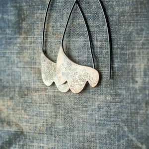 May include: A pair of silver earrings with a unique, organic shape and a textured finish. The earrings are suspended from black wire hoops.