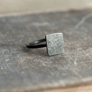Puede incluir: Un anillo de plata con una parte superior de forma cuadrada. La parte superior del anillo tiene un acabado texturizado y martillado.