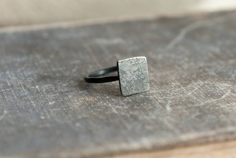 Puede incluir: Un anillo de plata con una parte superior de forma cuadrada. La parte superior del anillo tiene un acabado texturizado y martillado.
