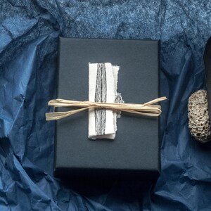 This photo shows the box the earrings will arrive in. It is a luxury black cardboard box with a hand drawn leaf design in ink. The box is shown with a black beach combed feather and a small stone which tell the story of the design‘s connection to nature. the background is dark blue. the box is tied with raffia for a rustic natural look.