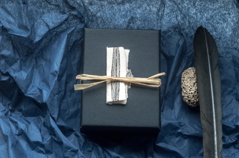 This photo shows the box the earrings will arrive in. It is a luxury black cardboard box with a hand drawn leaf design in ink. The box is shown with a black beach combed feather and a small stone which tell the story of the design‘s connection to nature. the background is dark blue. the box is tied with raffia for a rustic natural look.