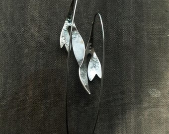 May include: A pair of silver-toned earrings with a leaf-like design. Each earring features three textured, elongated leaf shapes suspended from a thin, curved wire. The earrings are displayed against a dark, textured background.
