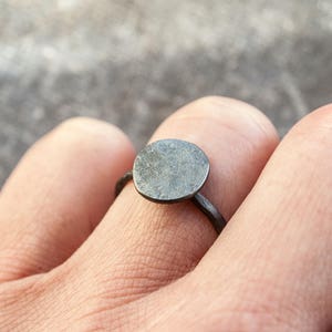 May include: A simple silver ring with a round, flat top. The ring is worn on a finger.