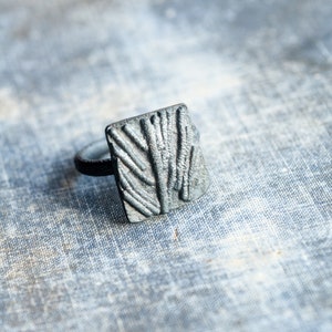 May include: A silver ring with a textured rectangular design. The ring is on a light blue fabric background.