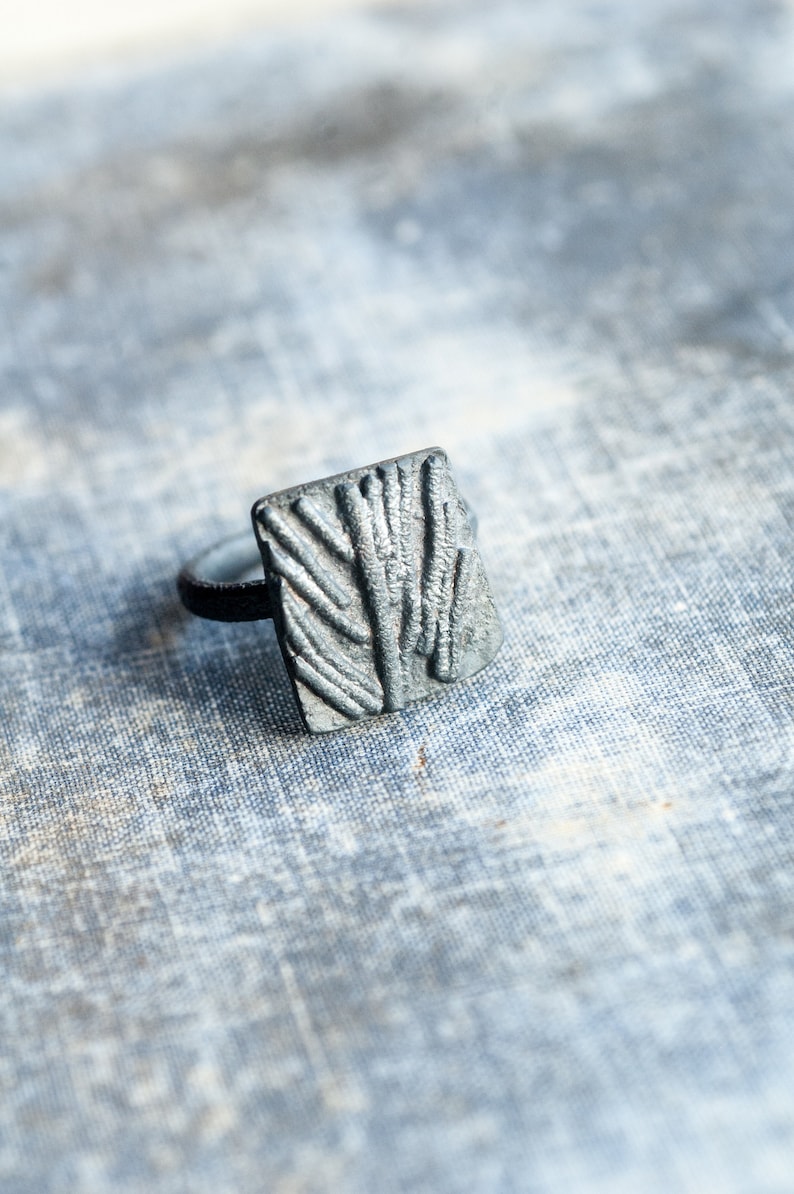 May include: A silver ring with a textured rectangular design. The ring is on a light blue fabric background.