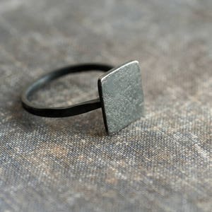 Puede incluir: Un anillo cuadrado negro con una superficie texturizada.