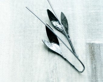 May include: Silver-toned leaf-shaped earrings with long, thin posts. The earrings have a textured surface and are arranged on a white, textured background. The design is minimalist and elegant, suitable for everyday wear or special occasions.