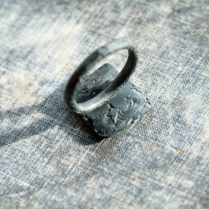 May include: A black metal ring with a textured surface. The ring is sitting on a gray fabric background.