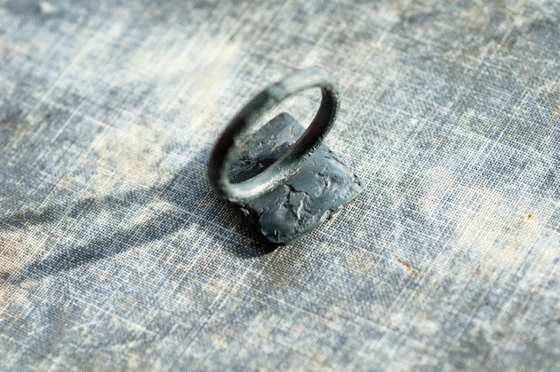 May include: A black metal ring with a textured surface. The ring is sitting on a gray fabric background.