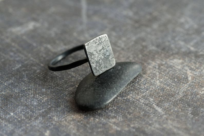 Puede incluir: Un anillo de plata con una parte superior de forma cuadrada. El anillo est&aacute; sobre una piedra negra y una superficie gris texturizada.