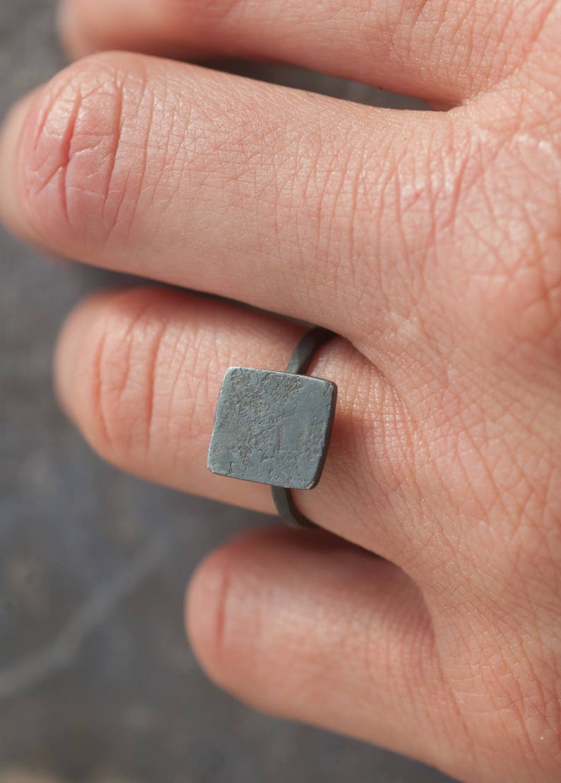 Puede incluir: Un anillo de forma cuadrada con un acabado texturizado gris oscuro. El anillo se lleva en un dedo.