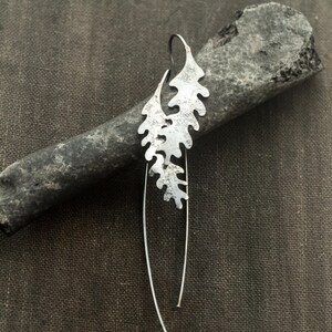 May include: A single silver earring with a leaf-like design. The earring features a textured, cutout pattern and a long, curved wire. The earring is displayed on a dark gray, textured surface.
