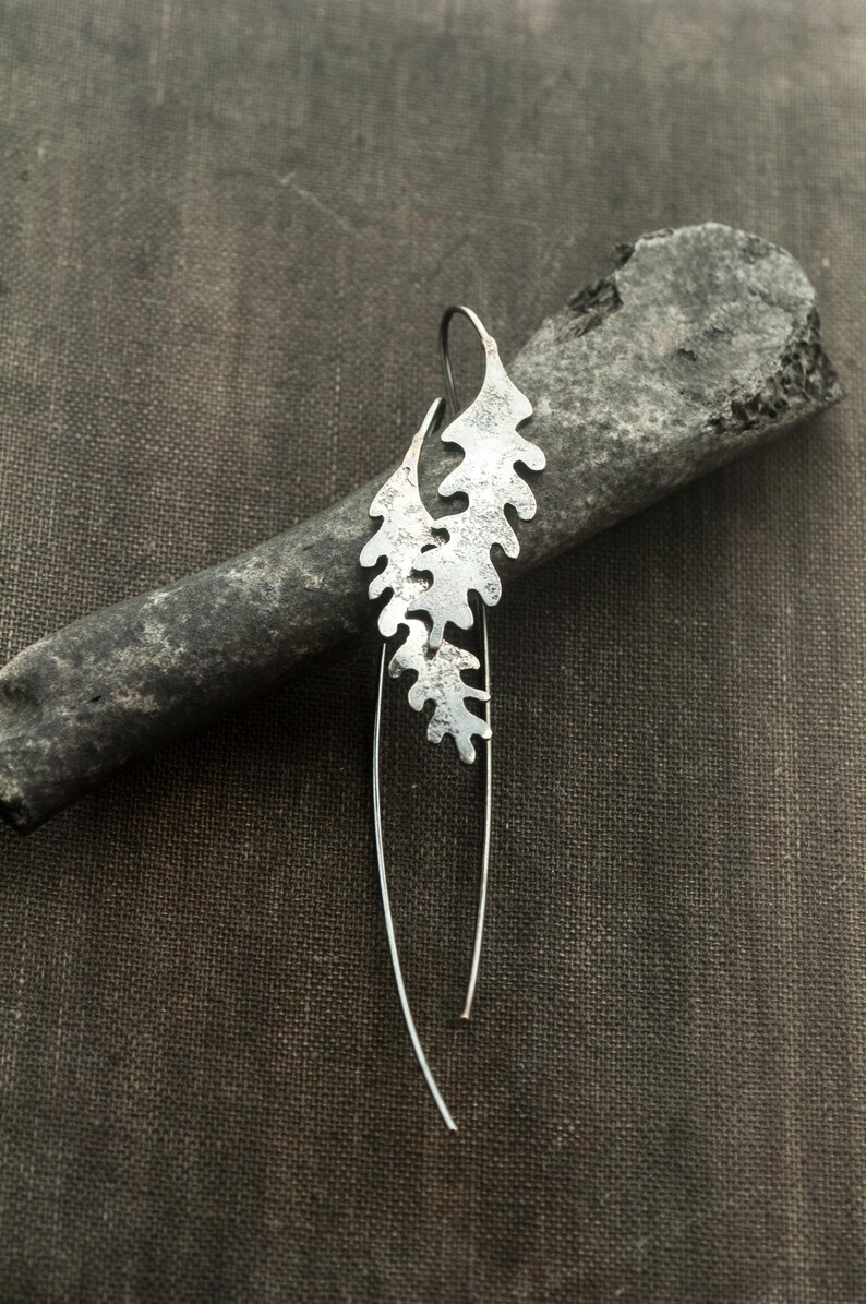 May include: A single silver earring with a leaf-like design. The earring features a textured, cutout pattern and a long, curved wire. The earring is displayed on a dark gray, textured surface.