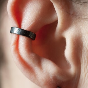Pode incluir: Close-up de uma orelha com um ear cuff preto e um pequeno brinco de prata. O ear cuff é uma faixa grossa com uma superfície texturizada. A pele é clara com um ligeiro tom rosado.