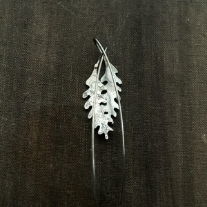 May include: A pair of silver-toned leaf-shaped earrings. Each earring features a detailed leaf design with a textured surface and long, slender wire accents. The earrings are set against a dark, textured background.