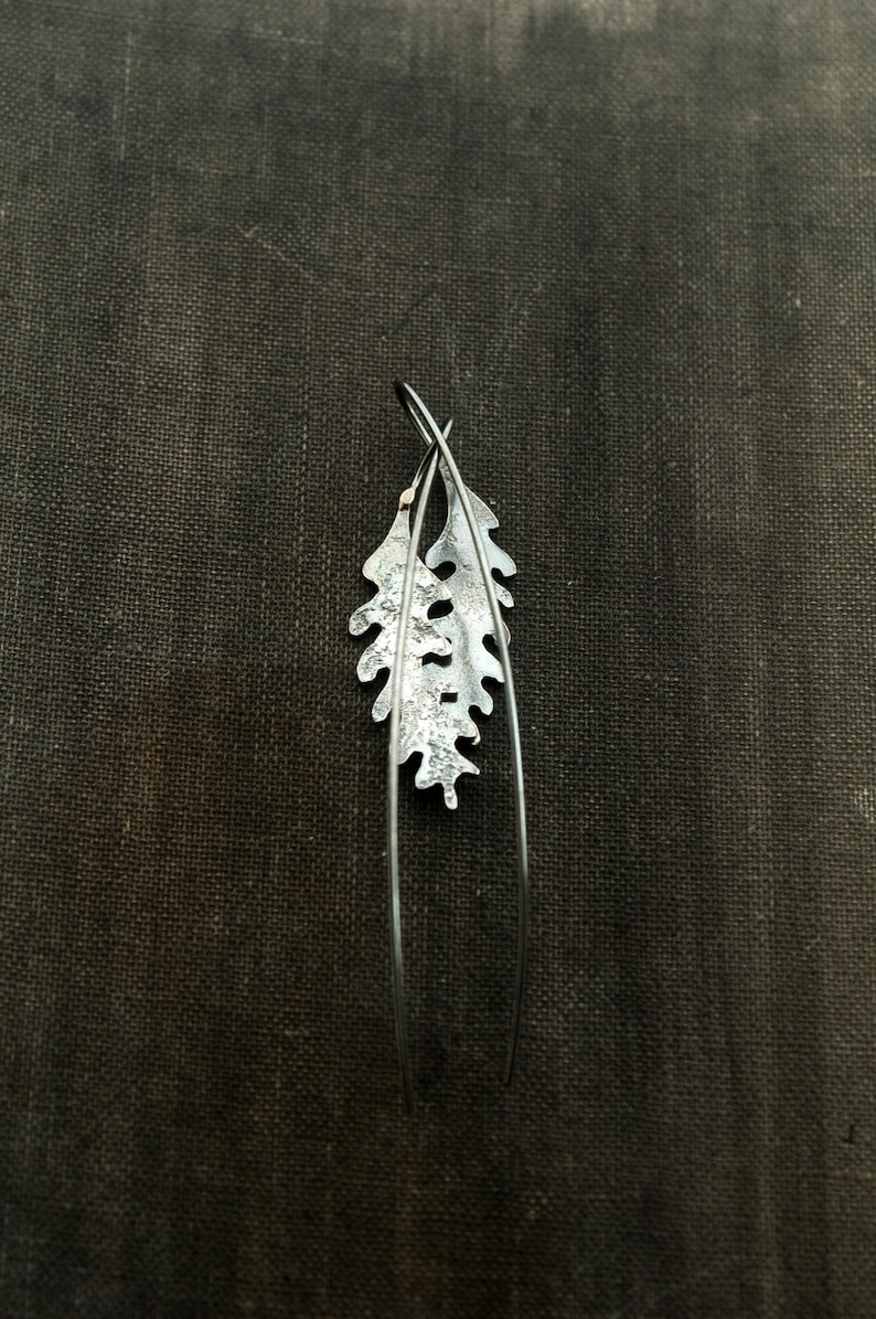 May include: A pair of silver-toned leaf-shaped earrings. Each earring features a detailed leaf design with a textured surface and long, slender wire accents. The earrings are set against a dark, textured background.