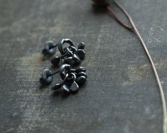 May include: A pair of dark gray stud earrings featuring a textured, organic, multi-layered circular design. Each earring consists of a central ring with several small, petal-like shapes dangling from it, secured with a matching dark gray post backing.