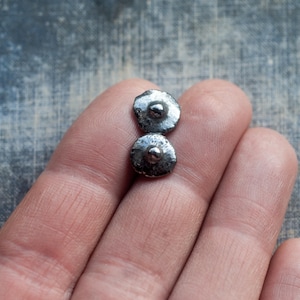 May include: Two small, silver-coloured, disc-shaped earrings with a raised centre are displayed. The earrings are held in a person's hand, showcasing their size and design. The background is a textured, muted blue surface.
