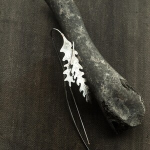 May include: A silver-colored earring with a leaf-like design, resting on a dark gray, textured stone. The earring has a long, thin wire that curves downward. The background is a dark, textured fabric.