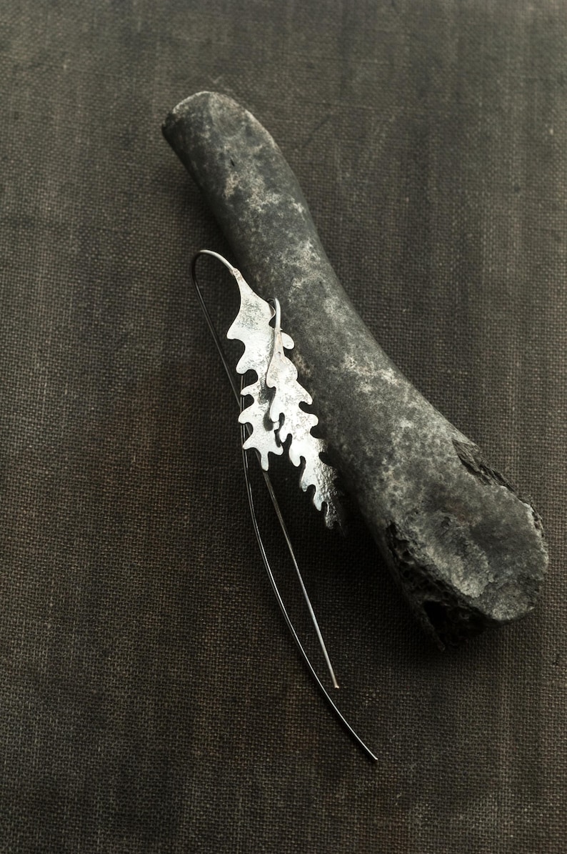 May include: A silver-colored earring with a leaf-like design, resting on a dark gray, textured stone. The earring has a long, thin wire that curves downward. The background is a dark, textured fabric.