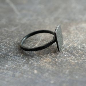 Puede incluir: Un anillo negro con una parte superior de forma cuadrada. El anillo est&aacute; hecho de metal y tiene un dise&ntilde;o simple.