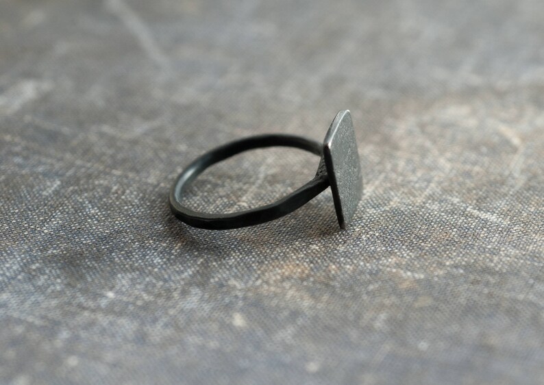 Puede incluir: Un anillo negro con una parte superior de forma cuadrada. El anillo est&aacute; hecho de metal y tiene un dise&ntilde;o simple.