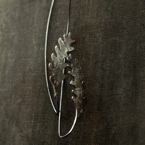 May include: A pair of silver-toned earrings with a botanical design. Each earring features a delicate, leaf-like shape with intricate detailing. The earrings hang from thin, curved wires, creating a graceful and natural aesthetic. The background is a dark, textured fabric.