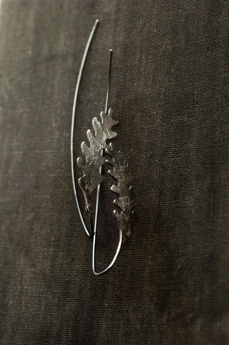 May include: A pair of silver-toned earrings with a botanical design. Each earring features a delicate, leaf-like shape with intricate detailing. The earrings hang from thin, curved wires, creating a graceful and natural aesthetic. The background is a dark, textured fabric.