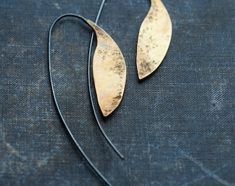 May include: A pair of gold leaf-shaped earrings with black wire hangers. The earrings have a hammered texture and a slightly irregular shape.