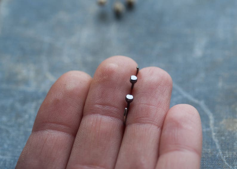 May include: Two silver stud earrings with square-shaped tops held in a hand.