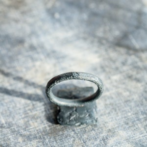May include: A dark gray metal ring with a textured surface. The ring is sitting on a gray textured surface.