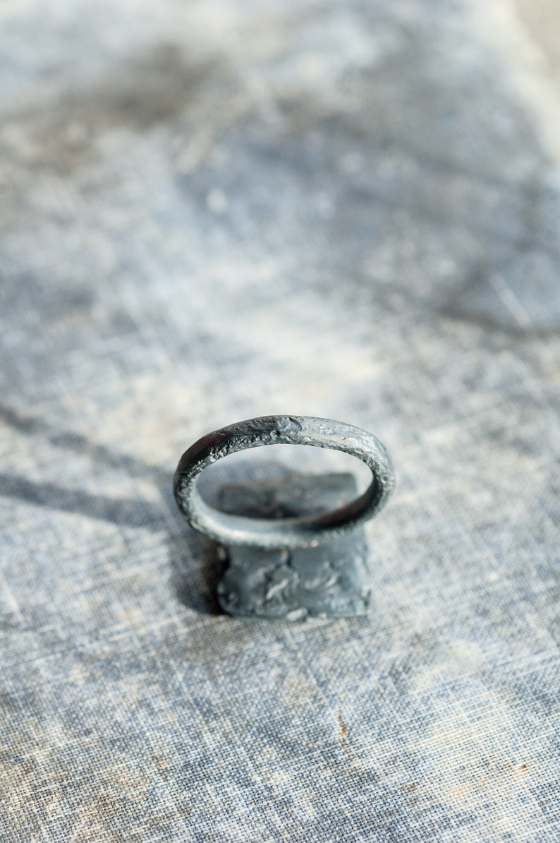 May include: A dark gray metal ring with a textured surface. The ring is sitting on a gray textured surface.