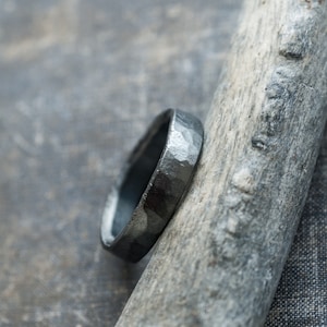 May include: A black hammered metal ring with a smooth, polished finish. The ring is displayed on a piece of driftwood.