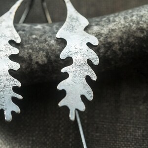 May include: Close-up of two silver-colored leaf-shaped earrings. The earrings have a textured surface and are suspended from thin silver wires. The background is a dark, textured surface.