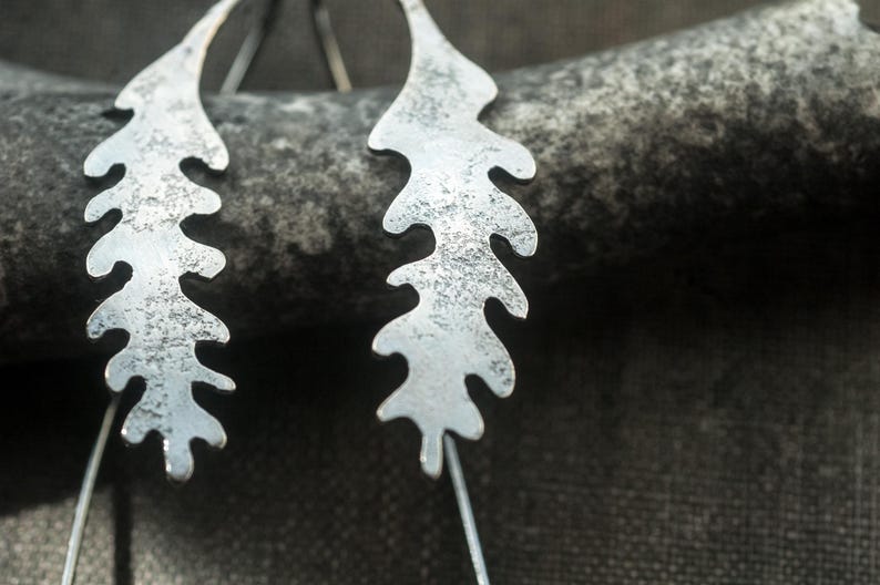 May include: Close-up of two silver-colored leaf-shaped earrings. The earrings have a textured surface and are suspended from thin silver wires. The background is a dark, textured surface.