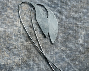 May include: A pair of silver leaf-shaped earrings with long, thin wires. The earrings are on a textured gray fabric background.