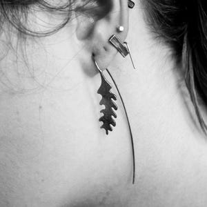 May include: Close-up of an ear adorned with multiple pieces of jewelry. A long, thin earring curves downward, while a leaf-shaped earring hangs from the earlobe. Other earrings include a small stud and a geometric bar.