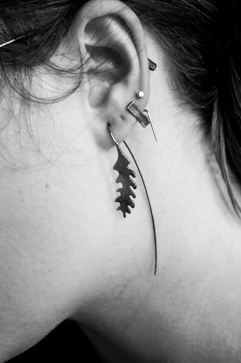 May include: Close-up of an ear adorned with multiple pieces of jewelry. A long, thin earring curves downward, while a leaf-shaped earring hangs from the earlobe. Other earrings include a small stud and a geometric bar.
