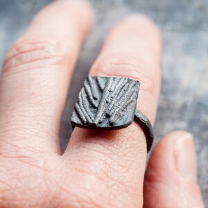 May include: A square-shaped ring with a textured design, made of dark metal. The ring is worn on a finger.
