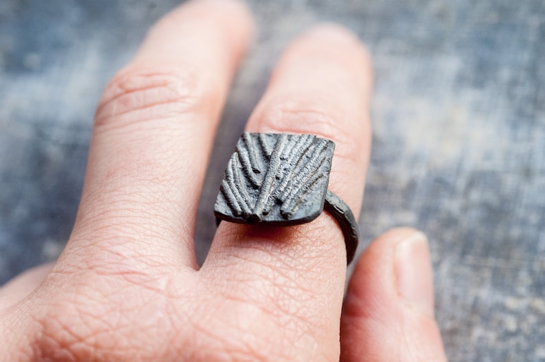 May include: A square-shaped ring with a textured design, made of dark metal. The ring is worn on a finger.