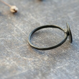 May include: A simple black ring with a round, flat, black stone. The ring is lying on a textured, grey fabric surface.