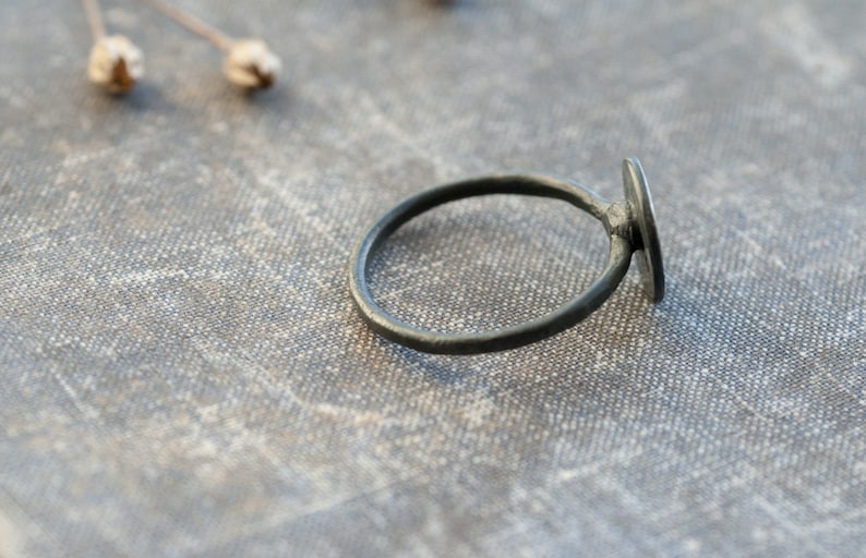 May include: A simple black ring with a round, flat, black stone. The ring is lying on a textured, grey fabric surface.