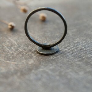May include: A black metal ring with a round, flat base. The ring is sitting on a textured, light brown surface.