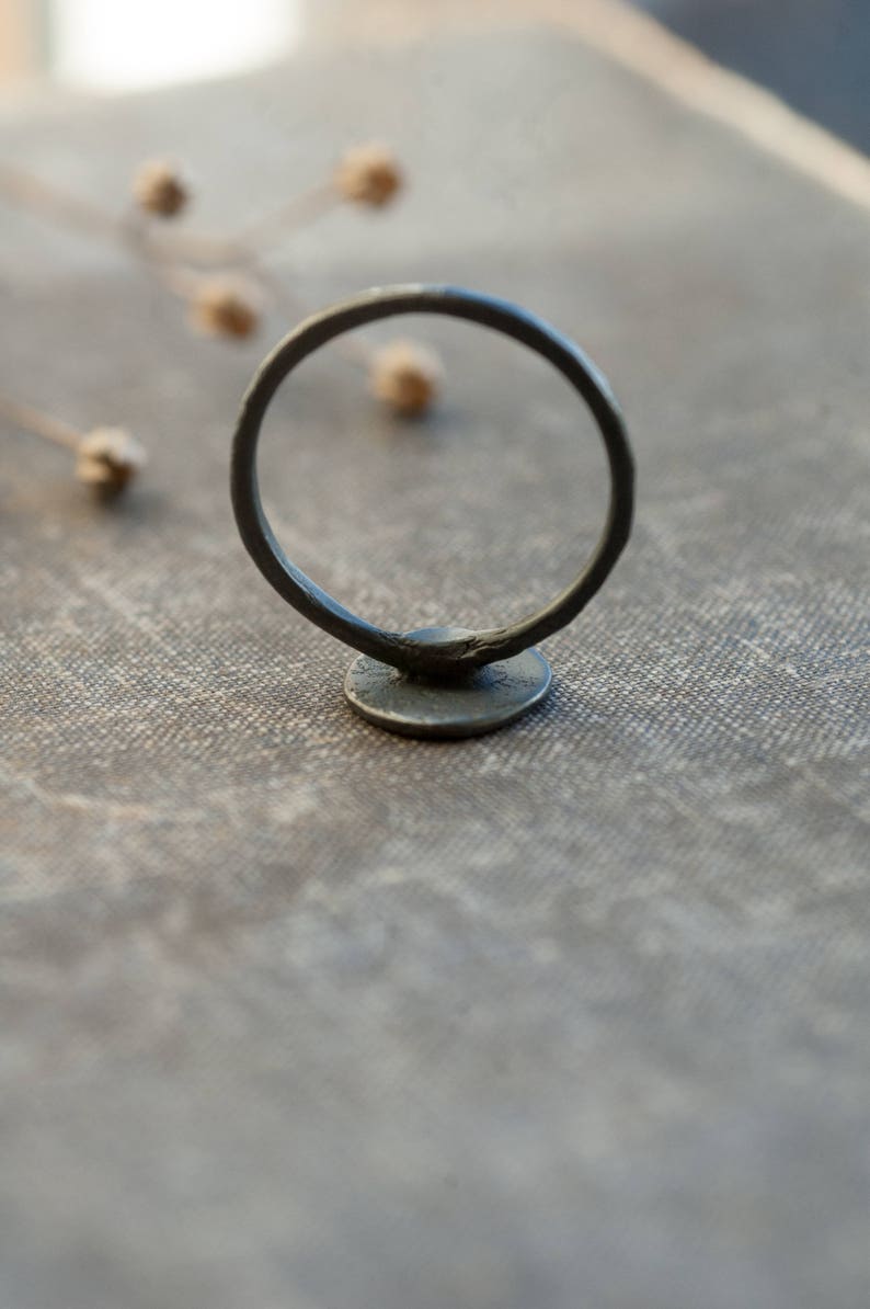 May include: A black metal ring with a round, flat base. The ring is sitting on a textured, light brown surface.