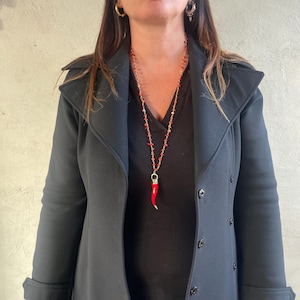 Può includere: Una lunga collana rossa con piccole perline rosse e una chiusura color oro. Il ciondolo è un peperoncino rosso. La collana è indossata sopra una camicia nera e un cappotto nero.