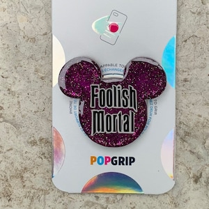 Puede incluir: Una agarradera para teléfono de purpurina morada con un diseño de cabeza de Mickey Mouse negro. La agarradera dice "Foolish Mortal" en letras negras. La agarradera está empaquetada en una tarjeta blanca con el logotipo de PopSockets.