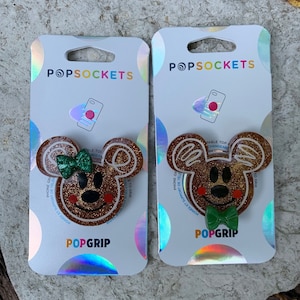 Może przedstawiać: Dwa uchwyty do telefonu PopSockets przedstawiające brązową i złotą głowę Myszki Miki z zielonym krawatem. Opakowanie jest srebrne z holograficznymi akcentami i tekstem "POPSOCKETS" i "POPGRIP".