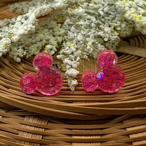 May include: A pair of pink glitter Mickey Mouse head earrings. The earrings are on a woven wicker surface.