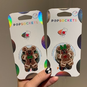 Puede incluir: Dos PopSockets para teléfono con un diseño de hombre de jengibre con detalles rojos y verdes. El hombre de jengibre lleva una corbata de lazo verde. El embalaje es plateado con detalles holográficos y el texto "PopSockets" y "PopGrip" impreso en él.