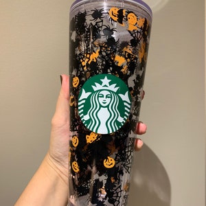 Op de afbeelding: Een doorzichtige plastic beker met een zwart en oranje Halloween-ontwerp. De beker heeft een Starbucks-logo, spinnen, spinnenwebben en pompoenen.